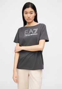 Szara koszulka z krótkim rękawem z nadrukowanym białym logo "EA7 EMPORIO ARMANI". Okrągły dekolt, luźny krój, gładka bawełniana faktura.
