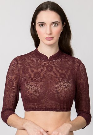 Frau trägt ein burgunderfarbenes, kurzes Spitzenoberteil mit dreiviertel Ärmeln, steht vor einem einfarbigen hellgrauen Hintergrund.