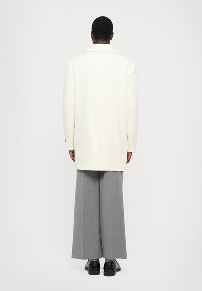 Cappotto bianco su misura con colletto a punta, abbinato a pantaloni grigi a gamba larga e scarpe nere, visto da dietro. Texture liscia.