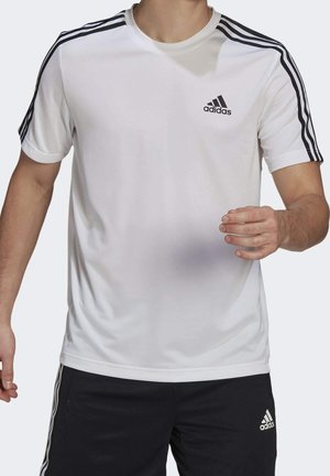 Homme portant un t-shirt blanc Adidas avec des bandes noires sur les épaules et un short Adidas noir, en mouvement avec les bras légèrement levés.