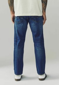 Vaqueros de mezclilla en azul oscuro, con un diseño clásico de pierna recta, textura sutilmente desvanecida y dos bolsillos traseros. Se llevan zapatillas blancas.