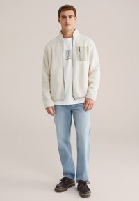 Chaqueta de forro polar color crema con cuello alto, cierre de cremallera y bolsillo en el pecho. Lucida sobre una camiseta gráfica blanca y jeans azul claro.