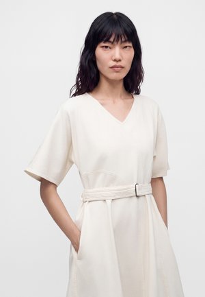 Femme aux longs cheveux foncés portant une robe crème courte à manches courtes ceinturée, avec un col en V et les mains dans les poches, debout devant un fond uni.