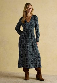 Langes, navyblaues Kleid mit weißem abstraktem Muster, V-Ausschnitt, langen Ärmeln, geraffter Taille und fließendem Rock, kombiniert mit braunen knielangen Stiefeln.