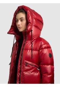 Nainen, joka käyttää kiiltävää punaista toppatakkia, jossa on suuri huppu, mustat vetoketjusomisteet ja musta logo hihoissa.