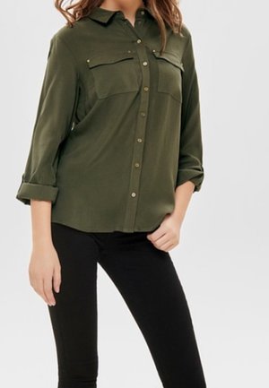 Femme portant une chemise boutonnée vert foncé avec des poches poitrine et des manches retroussées, associée à un pantalon noir, debout, une main sur la hanche.