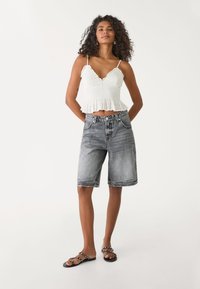 Top court smocké blanc avec des bords volantés, associé à un short bermuda en denim gris délavé. Les chaussures sont des sandales noires avec des clous décoratifs.