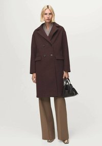 Cappotto in lana bordeaux doppiopetto con rever a punte, due tasche frontali e grandi bottoni neri, abbinato a pantaloni larghi marroni.
