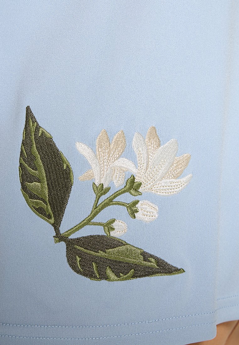 Jasnoniebieska tkanina z bogatym białym i zielonym haftem kwiatowym, zawierająca teksturowane liście i kwiaty, co dodaje wizualnego zainteresowania projektowi.