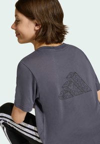 Sivý krátky rukáv tričko s geometrickým dizajnom loga na chrbte, s jemnou textúrou. Kombinované s čiernymi nohavicami so vzorovanými pruhmi.