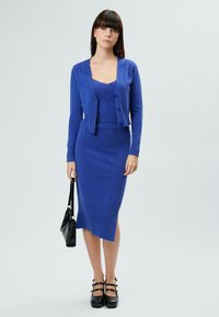 Blaues Strickkleid-Set mit einer Strickjacke und einem Rock, das über eine gerippte Textur und eine leicht taillierte Silhouette verfügt. Schwarze Handtasche und Plateauschuhe.