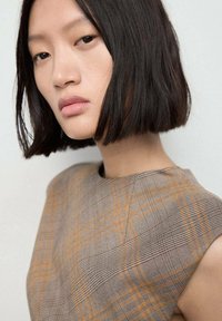 Bordeaux geruite mouwloze top met oranje accenten, textuurstof en een asymmetrische halslijn, die prachtig aansluit bij een rechte bob kapsel.