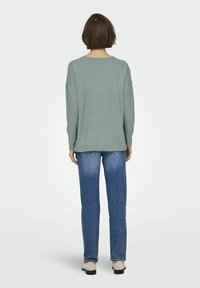 Groene langarmtrui van zachte stof, gecombineerd met lichtblauwe straight-leg jeans. Heeft een ontspannen pasvorm en verlaagde schouders.