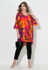 Farbenfrohe übergroße Tunika mit orangefarbenen, pinken und schwarzen abstrakten Mustern, kombiniert mit schwarzen Skinny-Hosen mit Nietenakzenten. Hält eine schwarze fransige Tasche.