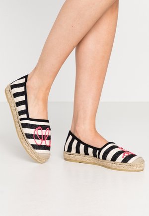 Ben iført sort-hvide stribede espadrillos med jutesåler og rødt broderet bogstav, stående på et lyst gulv.