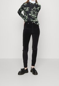 Svarta manchesterbyxor med hög midja och smal passform. Långärmad svart topp med ett grönt blommönster. Svarta skor med en dekorativ tofs.