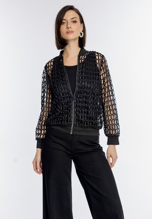 Kobieta ubrana w czarną kurtkę zapinaną na zamek z siateczkowym wzorem, na czarnym topie i czarnych spodniach, stojąca na tle jednolitego białego tła.