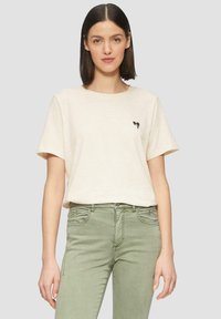 Beżowy bawełniany t-shirt z okrągłym dekoltem i krótkimi rękawami. Zawiera mały czarny nadruk serduszka na lewej piersi. Połączony z oliwkowymi spodniami.