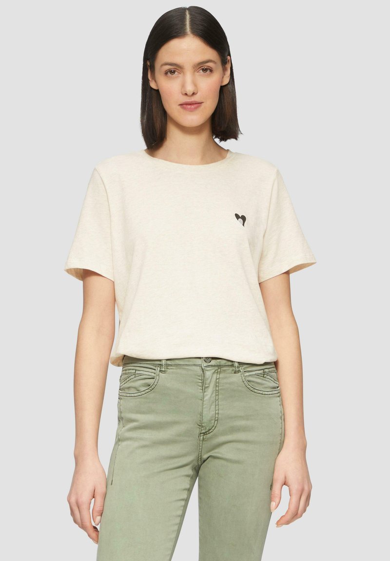 Beżowy bawełniany t-shirt z okrągłym dekoltem i krótkimi rękawami. Zawiera mały czarny nadruk serduszka na lewej piersi. Połączony z oliwkowymi spodniami.