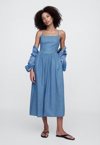 Traper haljina midi dužine sa kvadratnim izrezom, tankim naramenicama, nabranoj suknji i svijetloplavim pranjem. Uparena s traper jaknom i crnim sandalama.