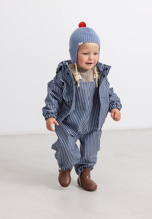 Tout-petit portant une combinaison de pluie bleu rayée, des bottes marron et un bonnet tricoté bleu avec un pompon rouge, marchant à l’intérieur sur un sol clair.