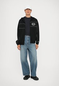 Crna zip-up dukserica s bijelim detaljima, mornarskoplavi T-shirt, svijetloplave široke traperice i crne cipele; model nosi bijelu kapu.