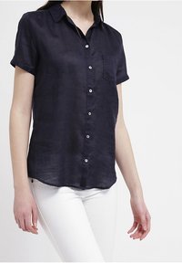 Námořnická modrá košile s krátkým rukávem na knoflíky, s límečkem, jednou náprsní kapsou a bílými knoflíky. Materiál vypadá lehký a hladký.