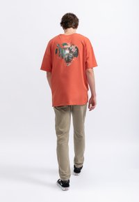 T-shirt arancione oversize con una grande grafica sulla schiena. Indossato con pantaloni beige chiaro e sneakers nere. Design semplice e dal taglio rilassato.
