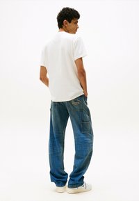 Camiseta blanca de algodón combinada con jeans de mezclilla azul de corte holgado, que presentan un bolsillo y bordado "Tommy" en la parte posterior. Zapatillas blancas completan el look.