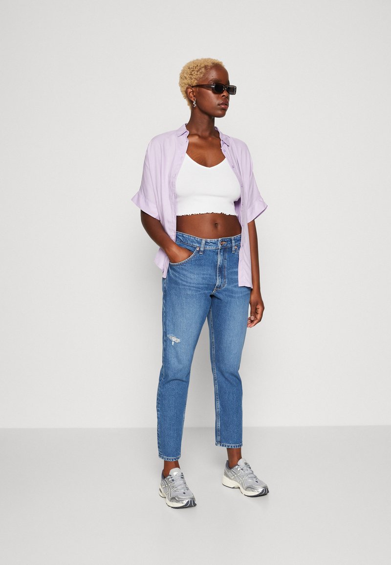 Hellblaues, kurzärmliges Hemd über einem weißen, taillierten Tanktop, kombiniert mit zerrissenen blauen Jeans und grauen Turnschuhen. DieAccessoires umfassen eine Sonnenbrille.