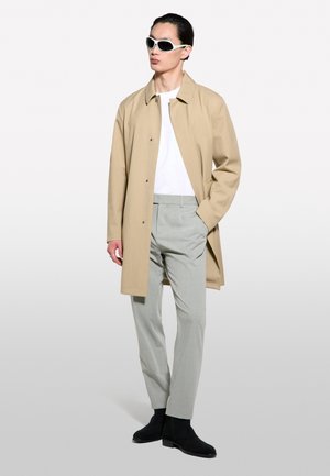 Homme portant un manteau beige, une chemise blanche, un pantalon gris clair, des bottes noires et des lunettes de soleil à monture blanche, debout avec une main dans la poche.