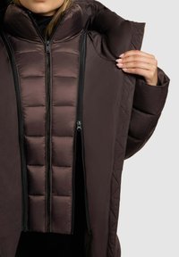 Brown padded jacket with a high collar, featuring a quilted texture and dual zippers on the front. Fabric is smooth with a matte finish.