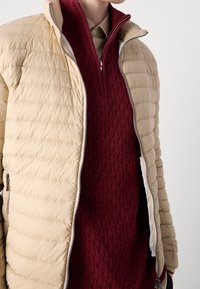 Beige pufferjacka med horisontella sömmar, med dragkedja framtill, bärs över en bordeauxfärgad stickad tröja med diamantmönster, lager på lager med en beige skjorta.