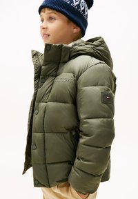 Veste matelassée vert olive avec capuche, présentant une texture matelassée, des boutons-pression et une patte de logo sur la manche.