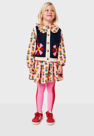 Colorful dress with playful patterns, navy knitted vest, festive collar, pink tights, red sneakers. Features floral design and button accents.