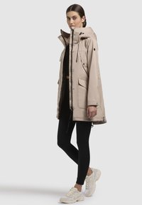 Parka impermeabile beige con cappuccio, zip frontale, bottoni a pressione e tasche laterali. Indossata sopra leggings neri e sneakers chiare.