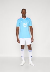 Camisola de futebol azul claro com mangas curtas, logótipo e detalhes brancos, combinada com calções brancos e meias brancas com riscas azuis.
