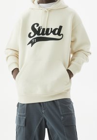 Personne portant un sweat à capuche crème avec le texte noir "Stud 91" et un pantalon cargo bleu marine, les mains dans les poches du sweat.