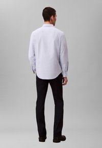 Chemise bleu clair à manches longues avec col boutonné, ourlet arrondi et poignets à un bouton, assortie à un pantalon foncé et des chaussures noires.