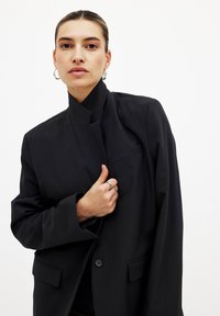 Blazer noir ajusté avec un col haut, fermeture à un bouton et poches avant. Tissu lisse avec une silhouette structurée.