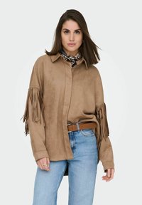 Brun ruskindsskjorte med frynser på ærmerne, knaplukning og krave. Kombineret med lyseblå jeans og et brunt bælte.