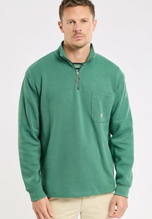 Homme portant un pull vert à demi-fermeture éclair avec poche poitrine, debout devant un fond blanc uni.