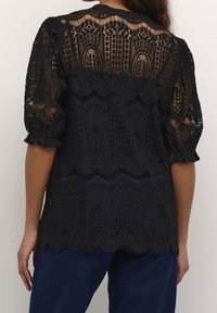 Blusa negra de encaje que presenta un diseño texturizado con mangas abullonadas y bordes con forma de olas, mostrando patrones intrincados y secciones transparentes.