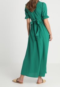 Vestido maxi verde con mangas cortas abullonadas, cinturón atado en la cintura, tejido fluido y textura suave. Combinado con sandalias planas.