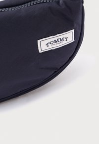 Námornícky nylonový vrecúško so zaobleným tvarom, ktoré má biely textilný štítok s nápisom "TOMMY HILFIGER" a zipsové zatváranie.