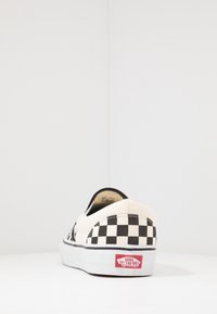 Zapatillas slip-on de lona con patrón de cuadros en blanco y negro, suela de goma blanca y acento del logo rojo de Vans en el talón.