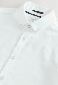 Bela srajca z gumbi iz teksturirane tkanine, značilna po ostrem ovratniku in šestih okroglih gumbih z logotipom.