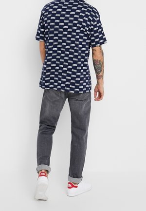 Mężczyzna w granatowej koszulce z logo Tommy Hilfiger, szarych dżinsach z podwiniętymi nogawkami, białych butach sportowych Adidas z czerwonymi detalami oraz z tatuażem na lewej ręce.