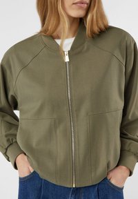 Kurtka typu bomber w kolorze oliwkowej zieleni, zapinana na zamek z przodu, z prążkowanymi mankietami i dwiema przednimi kieszeniami. Wykonana z miękkiej, teksturowanej tkaniny o luźnym kroju.