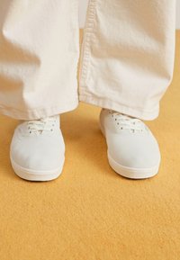 White canvas sneakers with round toe, flat laces, and rubber soles, worn with loose-fitting light-colored pants on a yellow background.